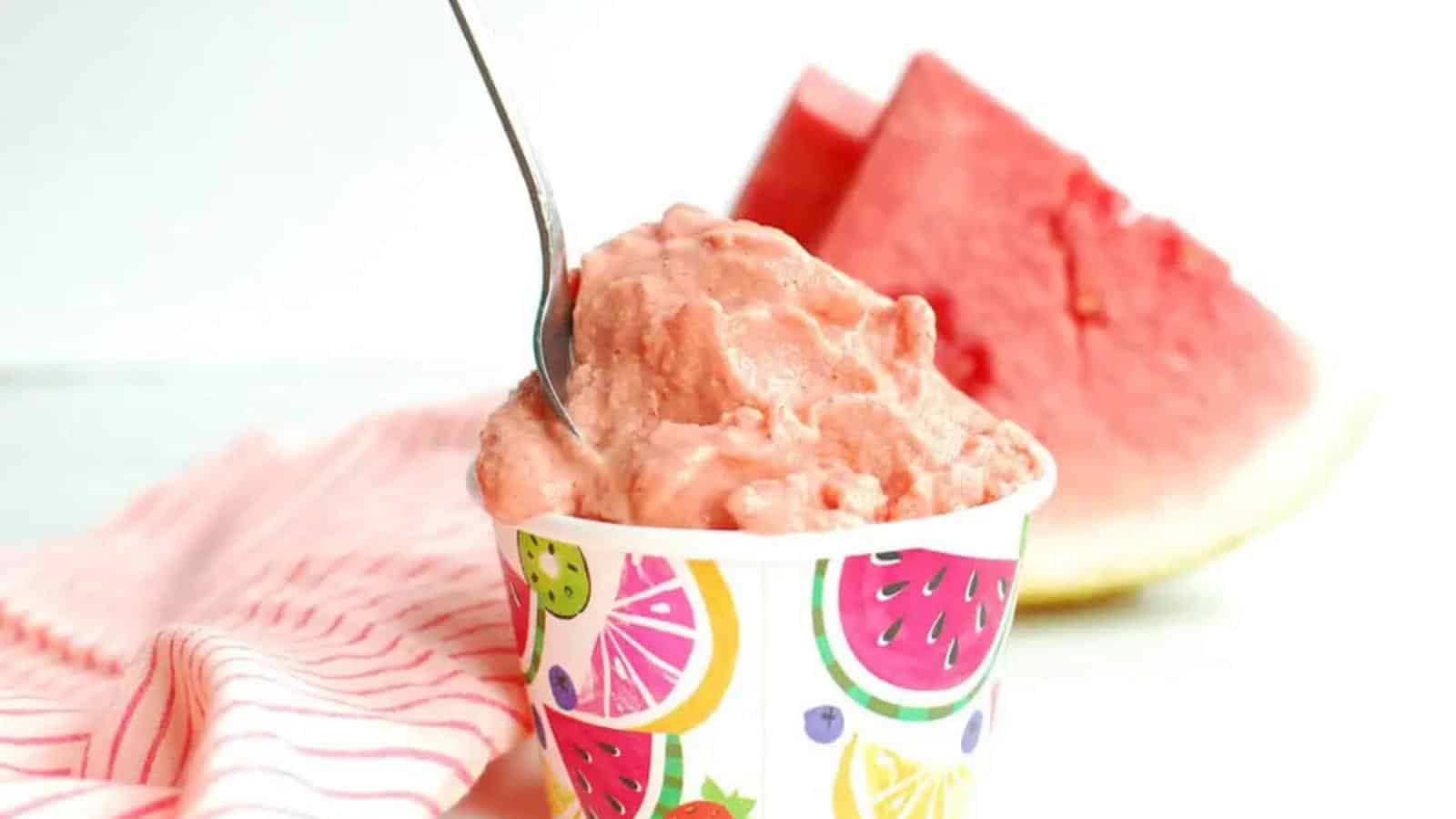 A cup of pink watermelon sorbet with a spoon, set on a striped cloth, with a slice of watermelon in the background.