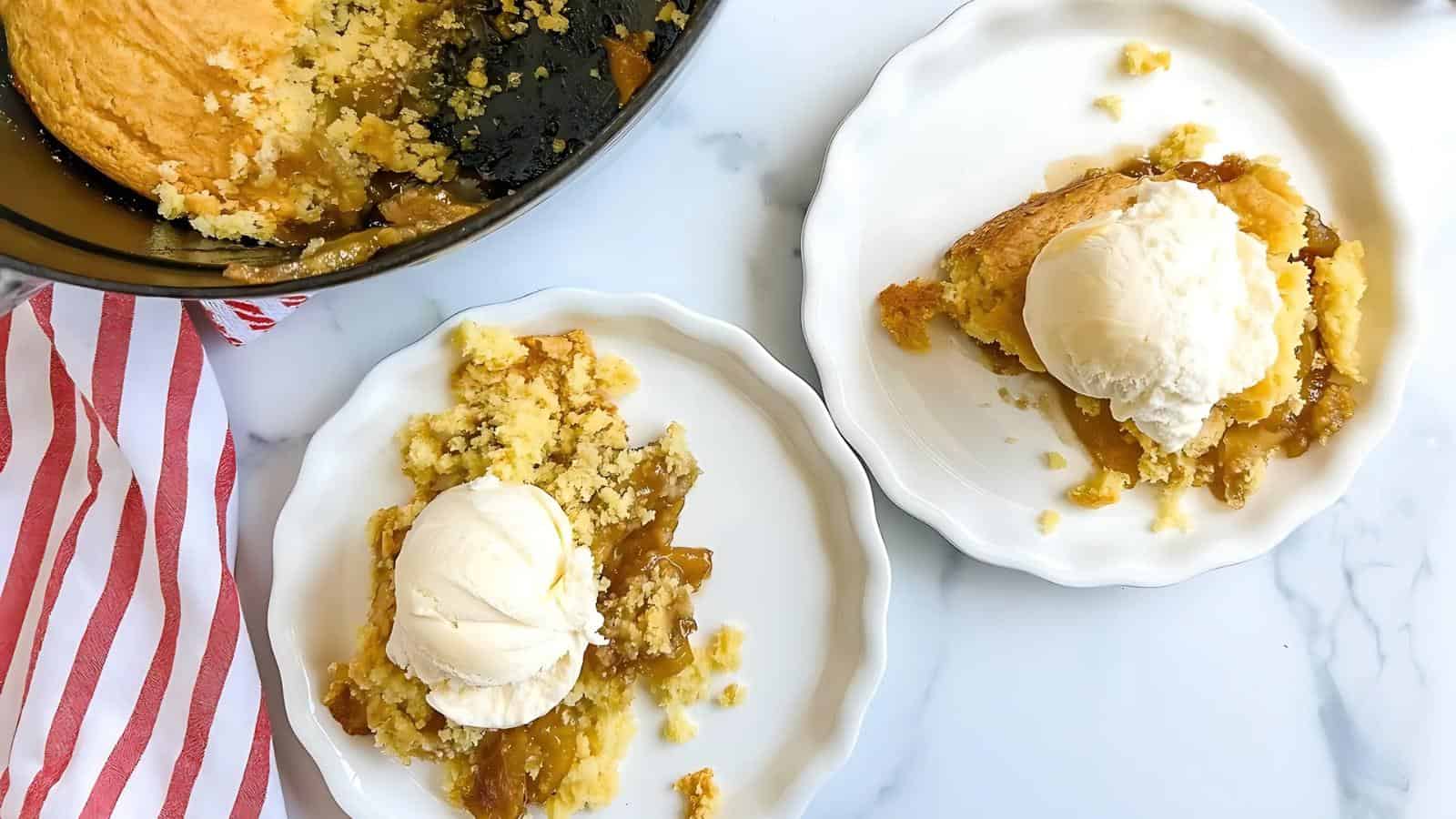 Two white plates with servings of fruit cobbler topped with vanilla ice cream, next to a baking dish with remaining cobbler and a red-striped napkin on a marble surface.