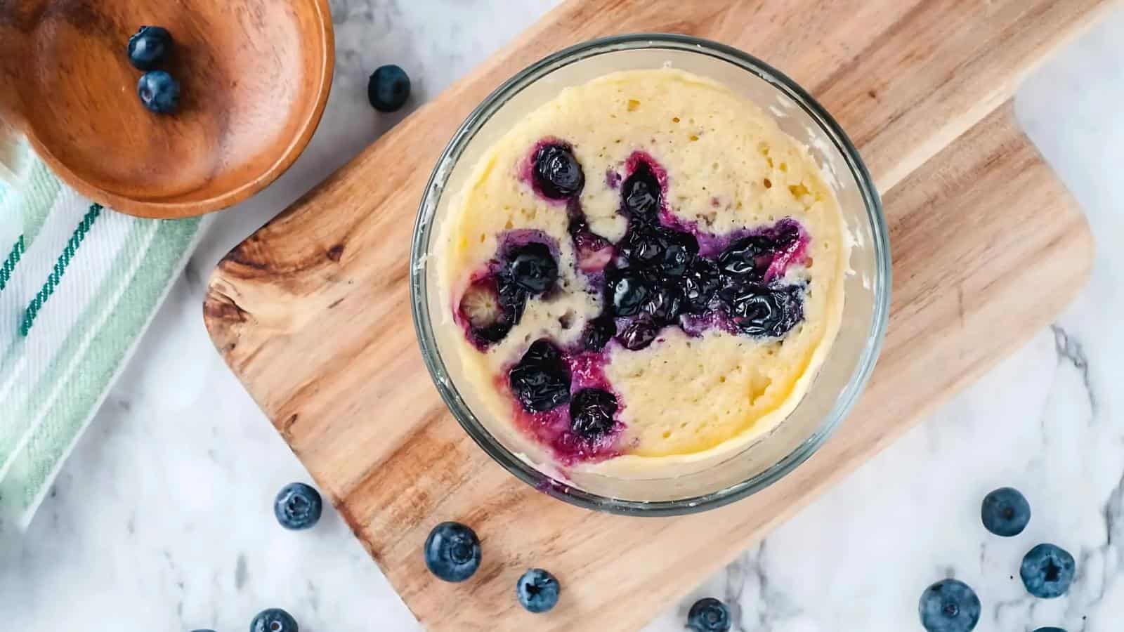 A glass bowl with a blueberry mug cake sits on a wooden board, surrounded by fresh blueberries and a wooden dish on a marble surface.