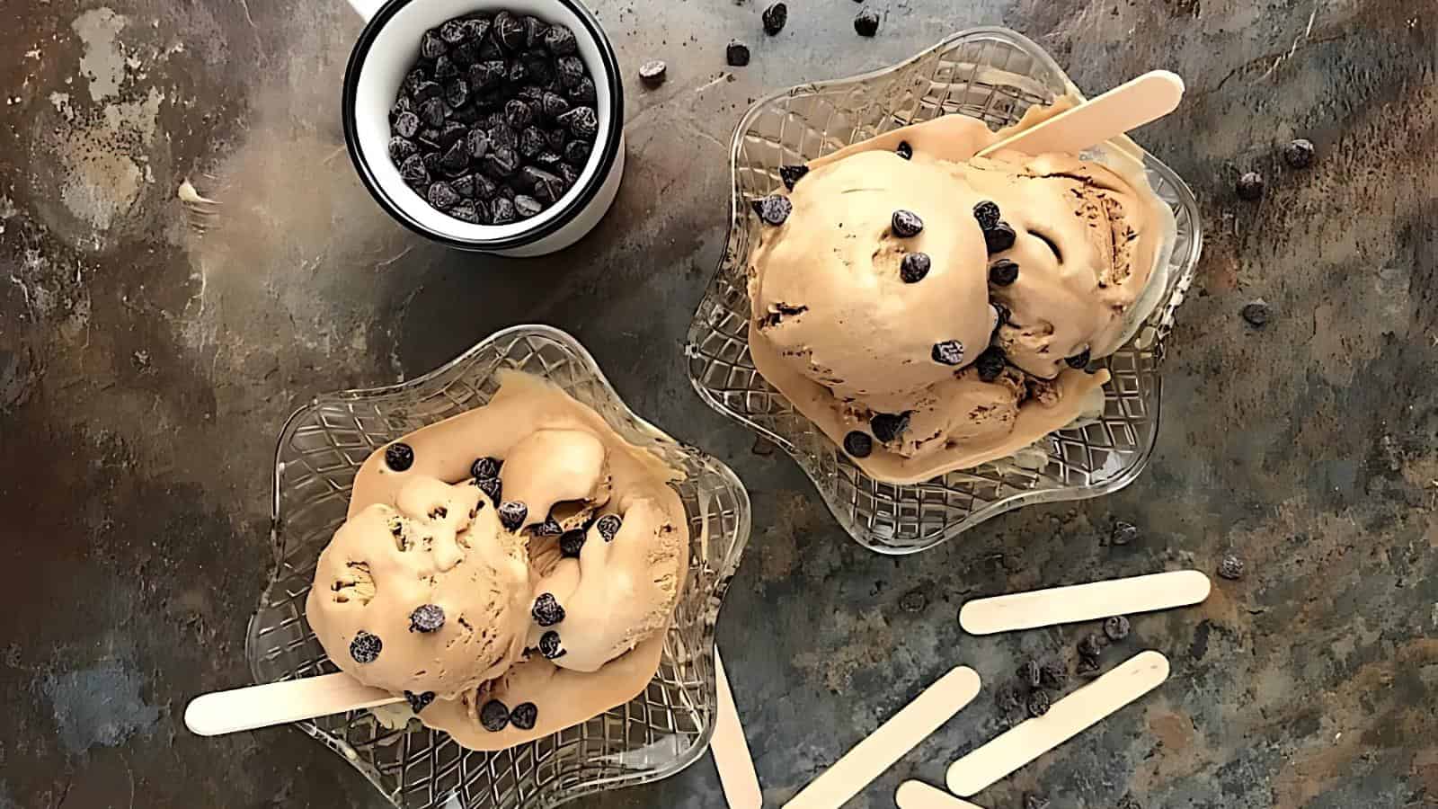 Two glass bowls of coffee ice cream with chocolate chips and wooden sticks, next to a small cup of chocolate chips and loose sticks on a textured surface.