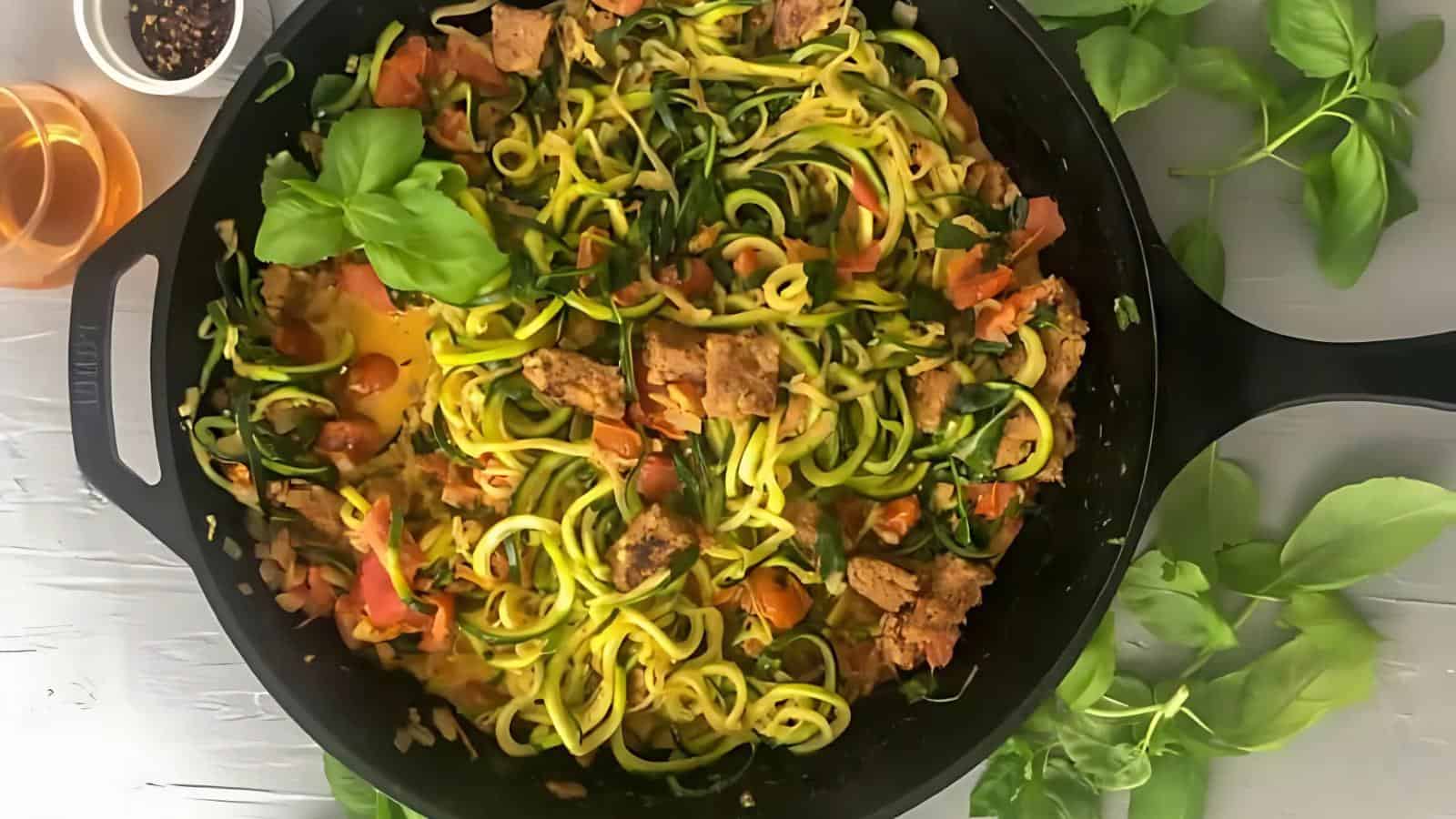 A skillet filled with zucchini noodles, diced tomatoes, pieces of cooked meat, and fresh basil, surrounded by basil leaves and a glass of light-colored liquid on a white surface.
