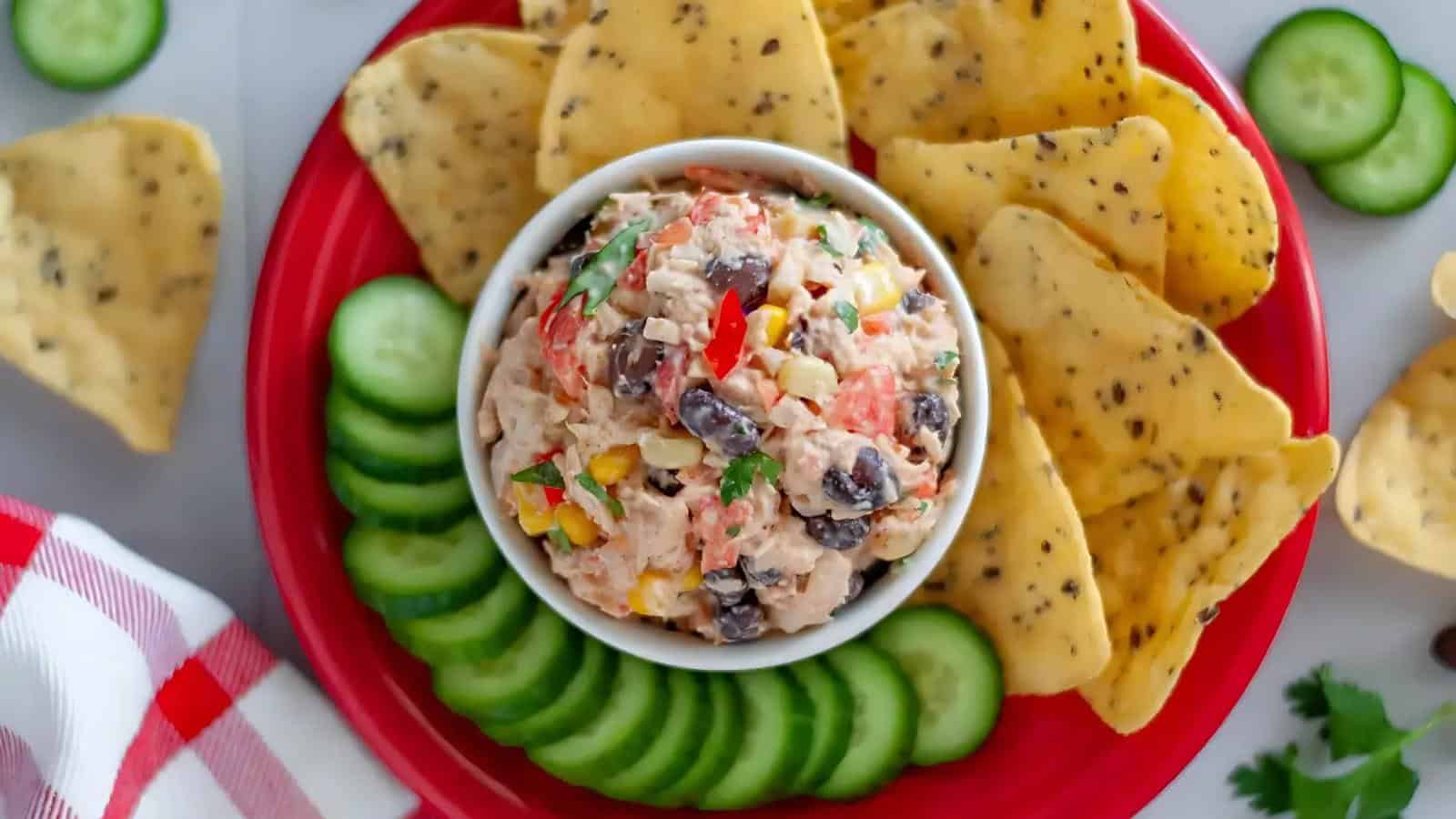 A bowl of mixed bean dip is surrounded by tortilla chips and cucumber slices on a red plate, with a red and white napkin nearby.