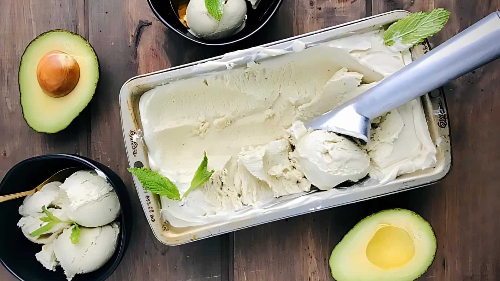 A tray of avocado ice cream with a scooper, two bowls of ice cream garnished with mint, and fresh avocado halves on a wooden surface.