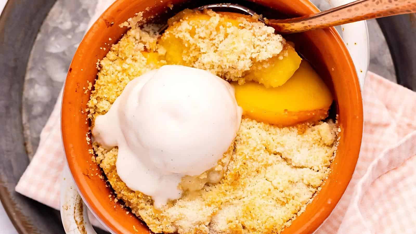 A baked peach crumble dessert in a small clay dish, topped with a scoop of vanilla ice cream and served with a spoon.
