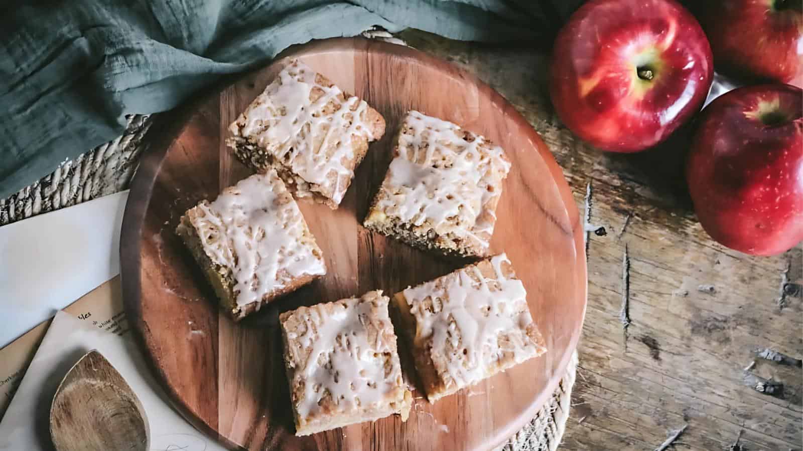 Five glazed apple cake bars are arranged on a round wooden board next to three red apples on a rustic wooden surface.