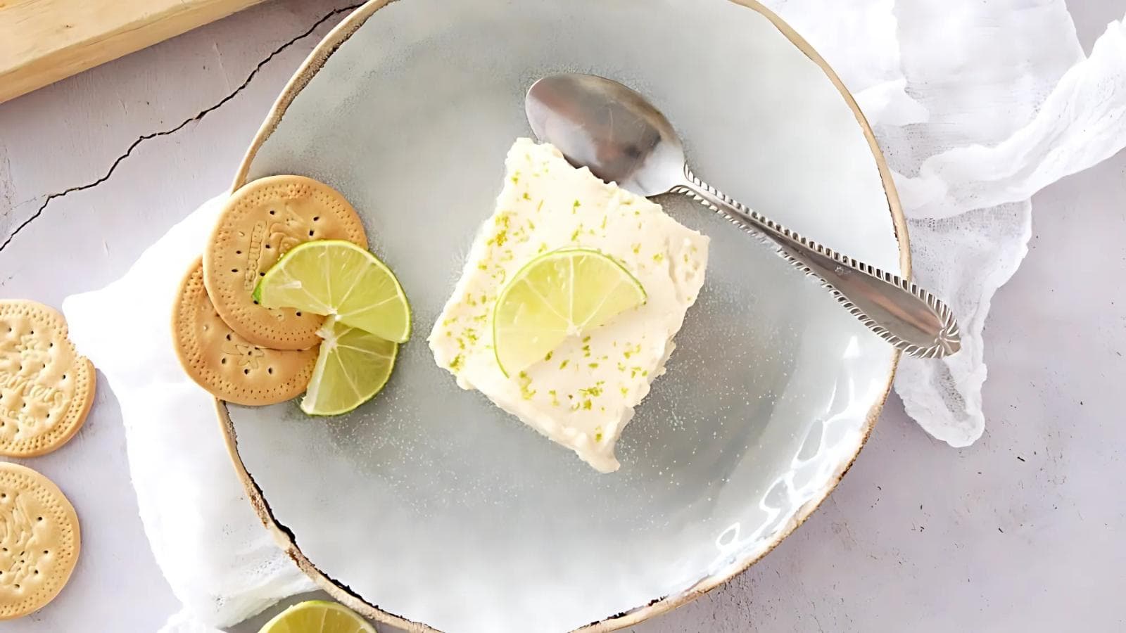 A square slice of lime cheesecake garnished with lime slices, served on a gray plate with a spoon and round crackers on the side.