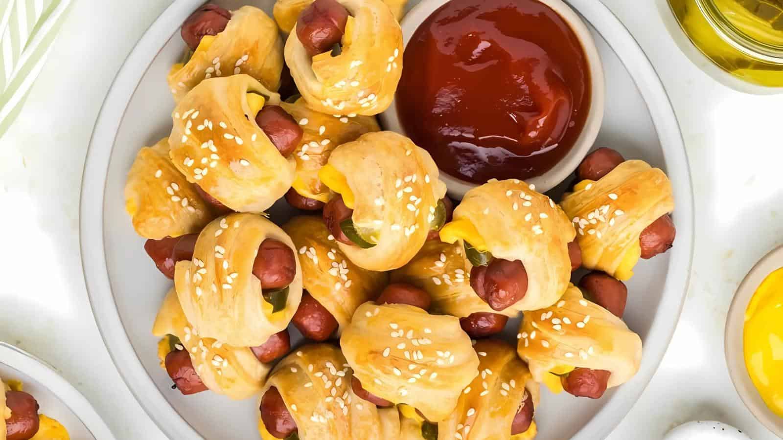 A plate of mini pigs in a blanket with sesame seeds, served with a bowl of ketchup for dipping.