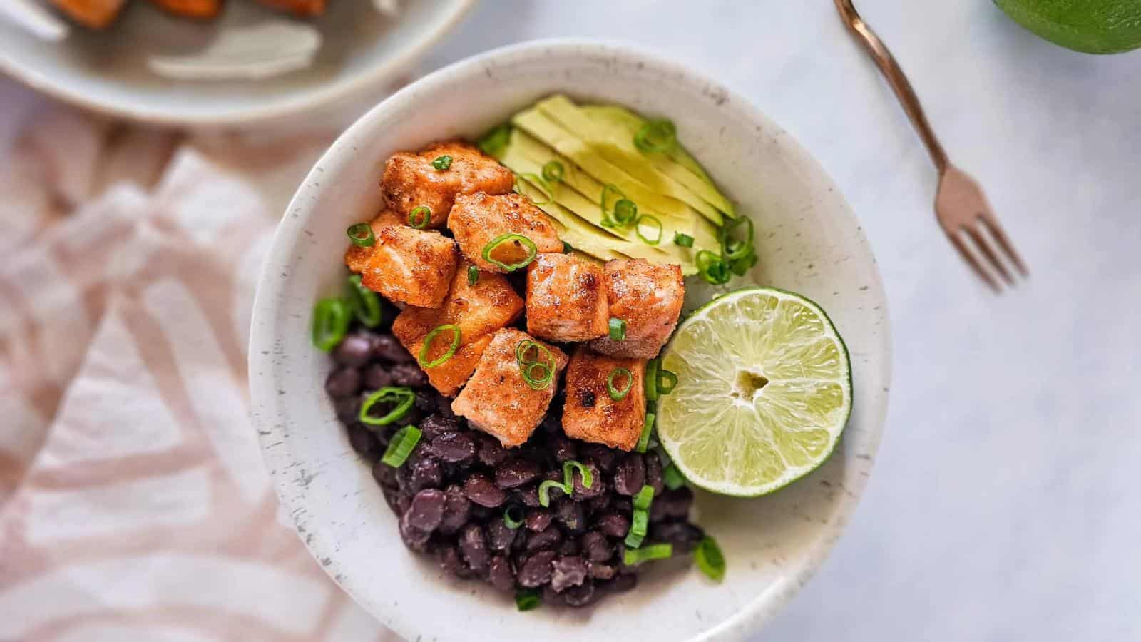 A bowl containing black beans, cubed seasoned tofu, sliced avocado, chopped green onions, and half a lime.