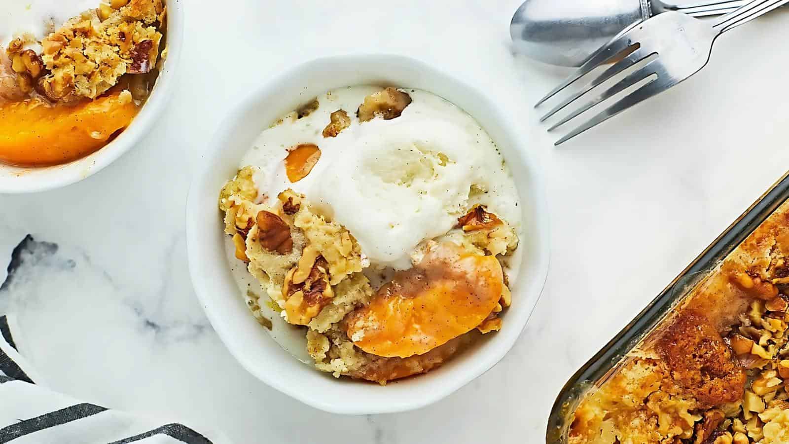 A bowl of peach cobbler topped with vanilla ice cream sits on a white surface, alongside a baking dish, utensils, and a second bowl of cobbler.