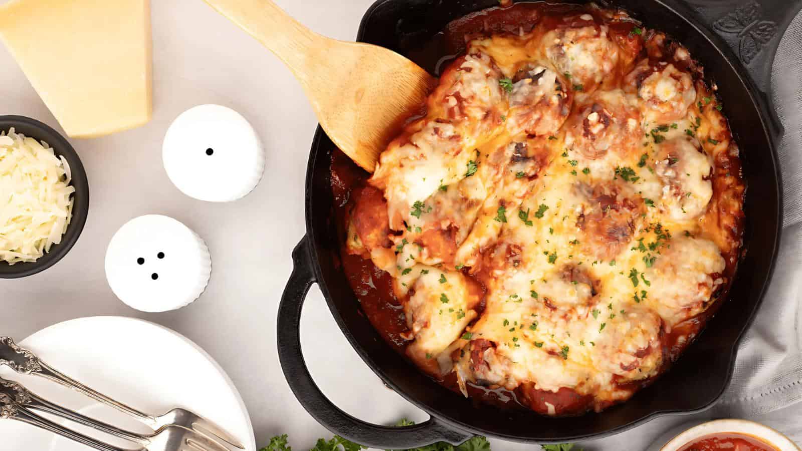 A cast iron skillet filled with baked meatballs in tomato sauce, topped with melted cheese and herbs, sits on a table beside plates, utensils, grated cheese, and salt and pepper shakers.