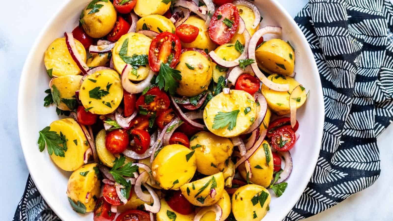 A bowl of potato salad with sliced yellow potatoes, cherry tomatoes, red onion, and parsley on a patterned napkin.