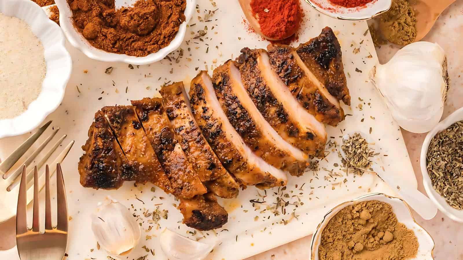 Sliced grilled chicken breast on a cutting board, surrounded by various spices in small bowls, garlic cloves, and utensils.