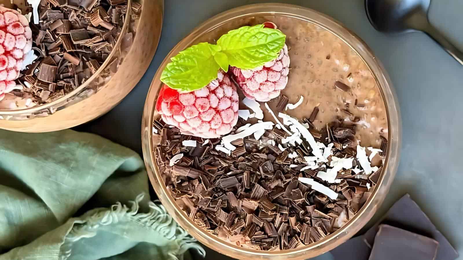 A glass bowl of chocolate chia pudding topped with frozen raspberries, mint leaves, shredded coconut, and chocolate shavings, with a spoon and green cloth nearby.