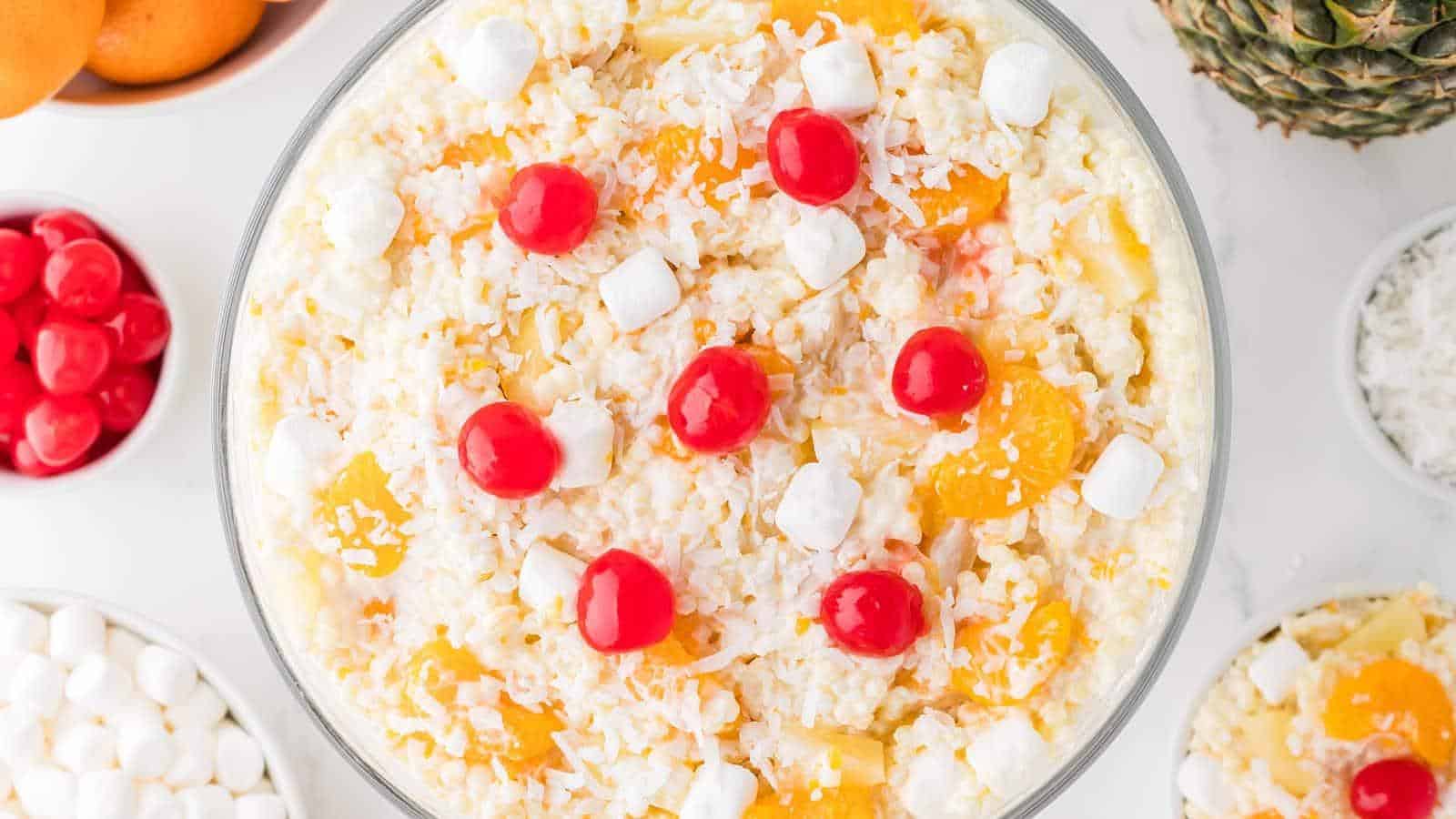 A glass bowl filled with a creamy fruit salad topped with maraschino cherries, mini marshmallows, shredded coconut, and fruit pieces.