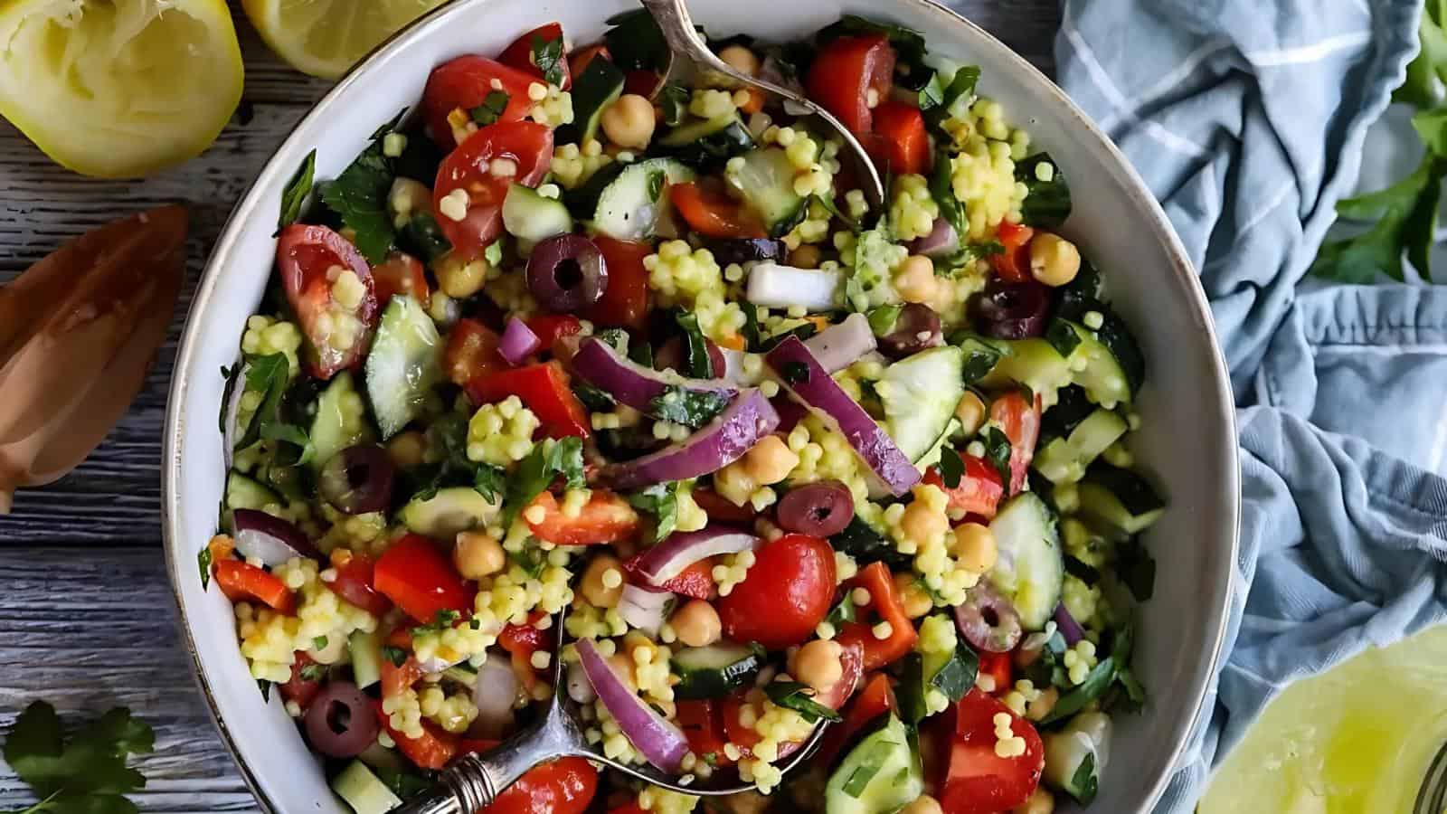 A bowl of Mediterranean salad with couscous, chickpeas, tomatoes, cucumbers, red onions, olives, and herbs, with serving utensils in the bowl.