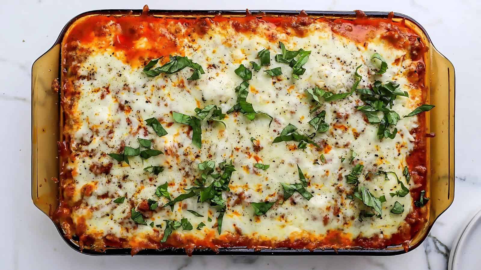 A baked lasagna in a rectangular glass dish, topped with melted cheese, tomato sauce, and fresh chopped basil.