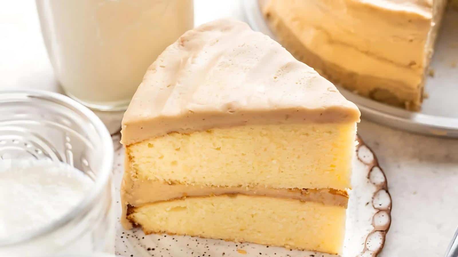 A slice of yellow layer cake with light brown frosting sits on a plate, with a glass of milk and part of the remaining cake visible in the background.