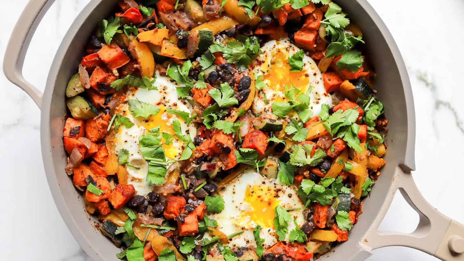 A skillet filled with baked eggs, black beans, diced sweet potatoes, bell peppers, onions, and cilantro garnish.