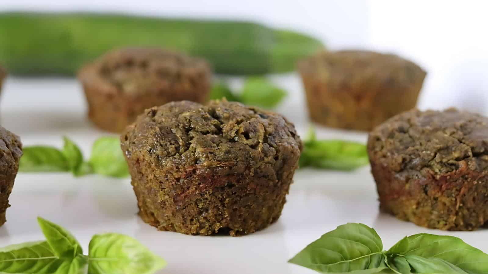 Several zucchini muffins are displayed on a white surface, surrounded by fresh basil leaves, with a whole zucchini in the background.