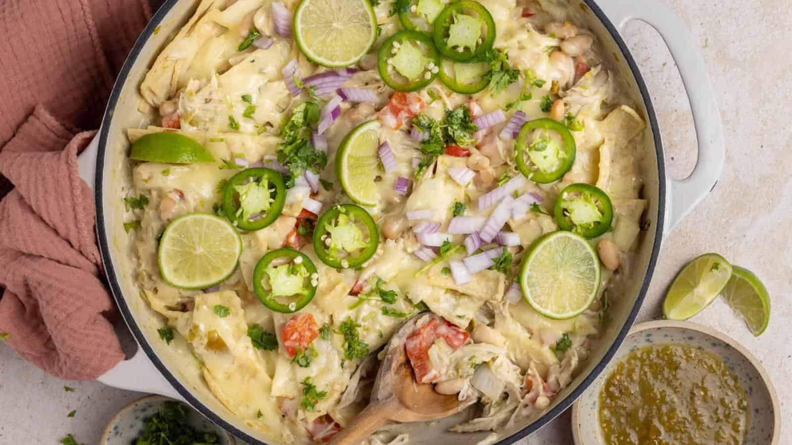 A creamy chicken casserole topped with sliced jalapeños, lime wedges, chopped red onions, and herbs in a round white dish, with a wooden spoon and a beige napkin beside it.
