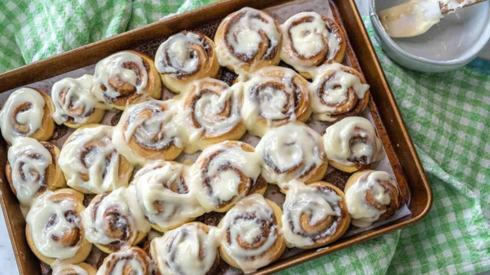 A baking tray filled with cinnamon rolls topped with white icing sits on a green checkered cloth.
