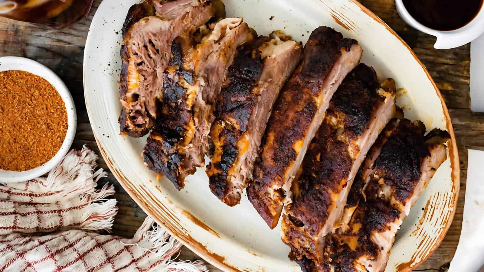 A platter of sliced, cooked pork ribs with a dark, seasoned crust is served on a white oval plate beside a red-and-white cloth.