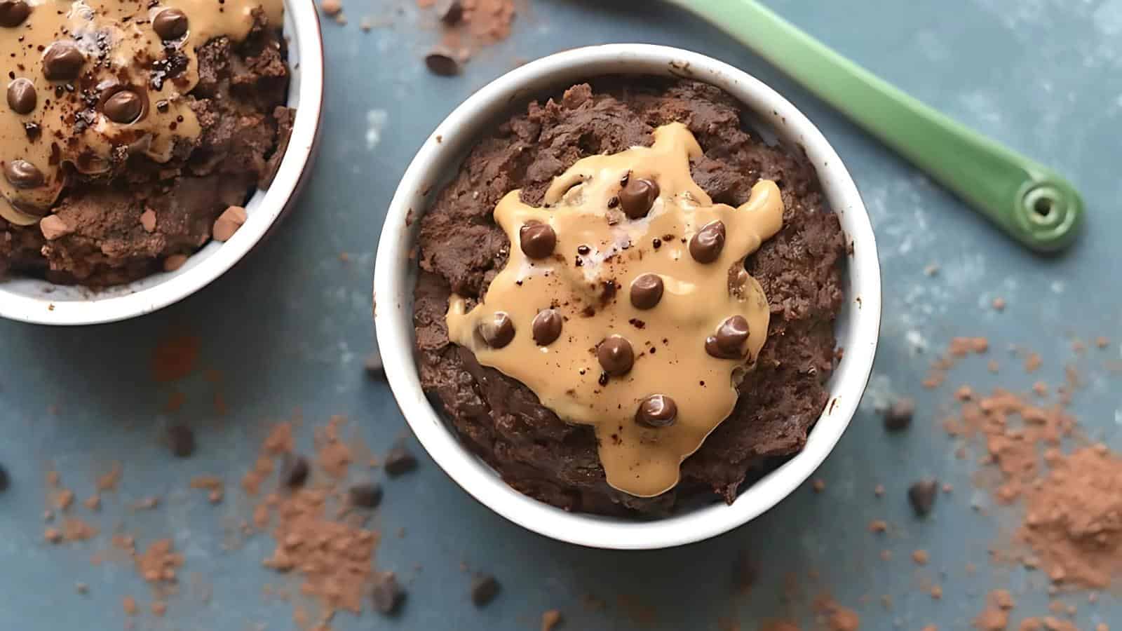 A white ramekin filled with chocolate mug cake topped with peanut butter and chocolate chips, placed on a blue surface with scattered cocoa powder and chocolate chips.