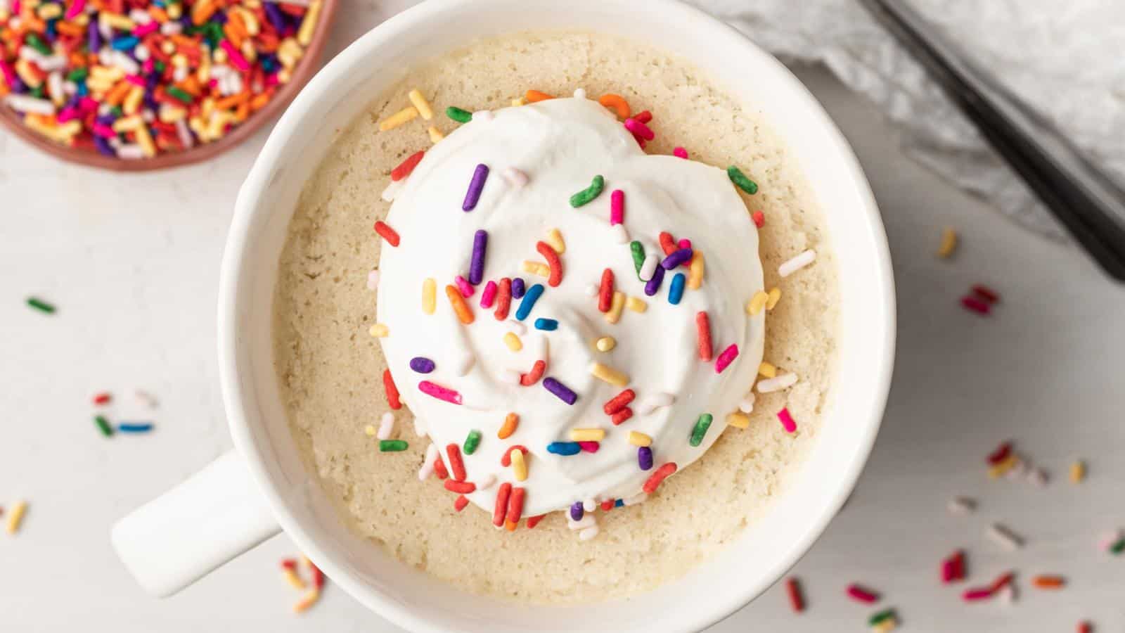 A mug cake topped with whipped cream and colorful sprinkles sits on a white surface, with extra sprinkles scattered nearby.