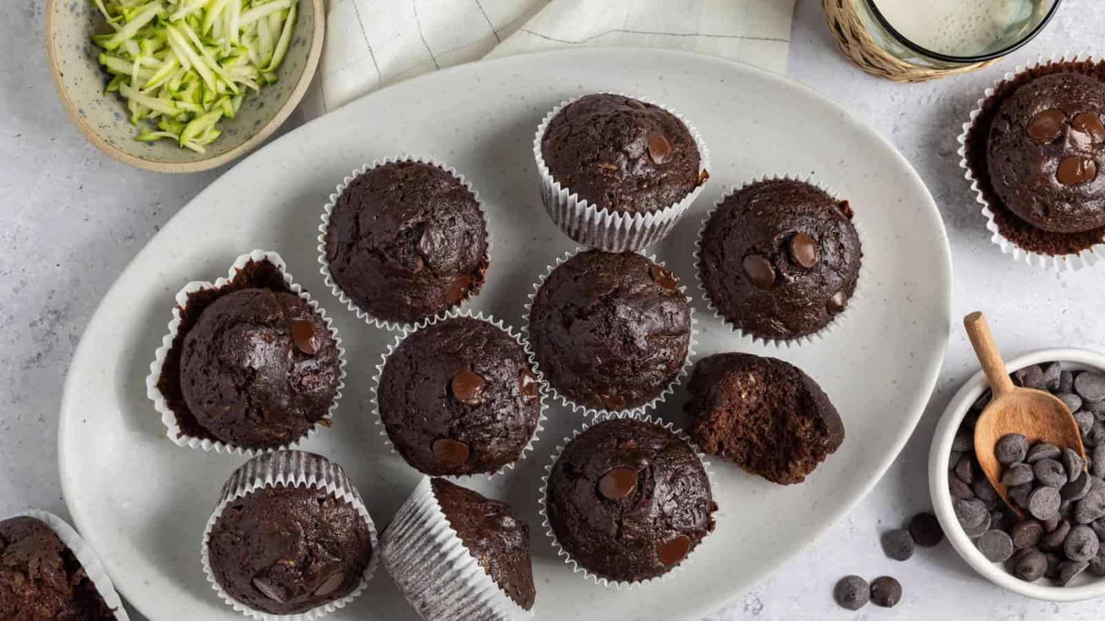 Chocolate muffins with chocolate chips are arranged on an oval plate, with zucchini strips, chocolate chips, and a glass of milk nearby on a light surface.