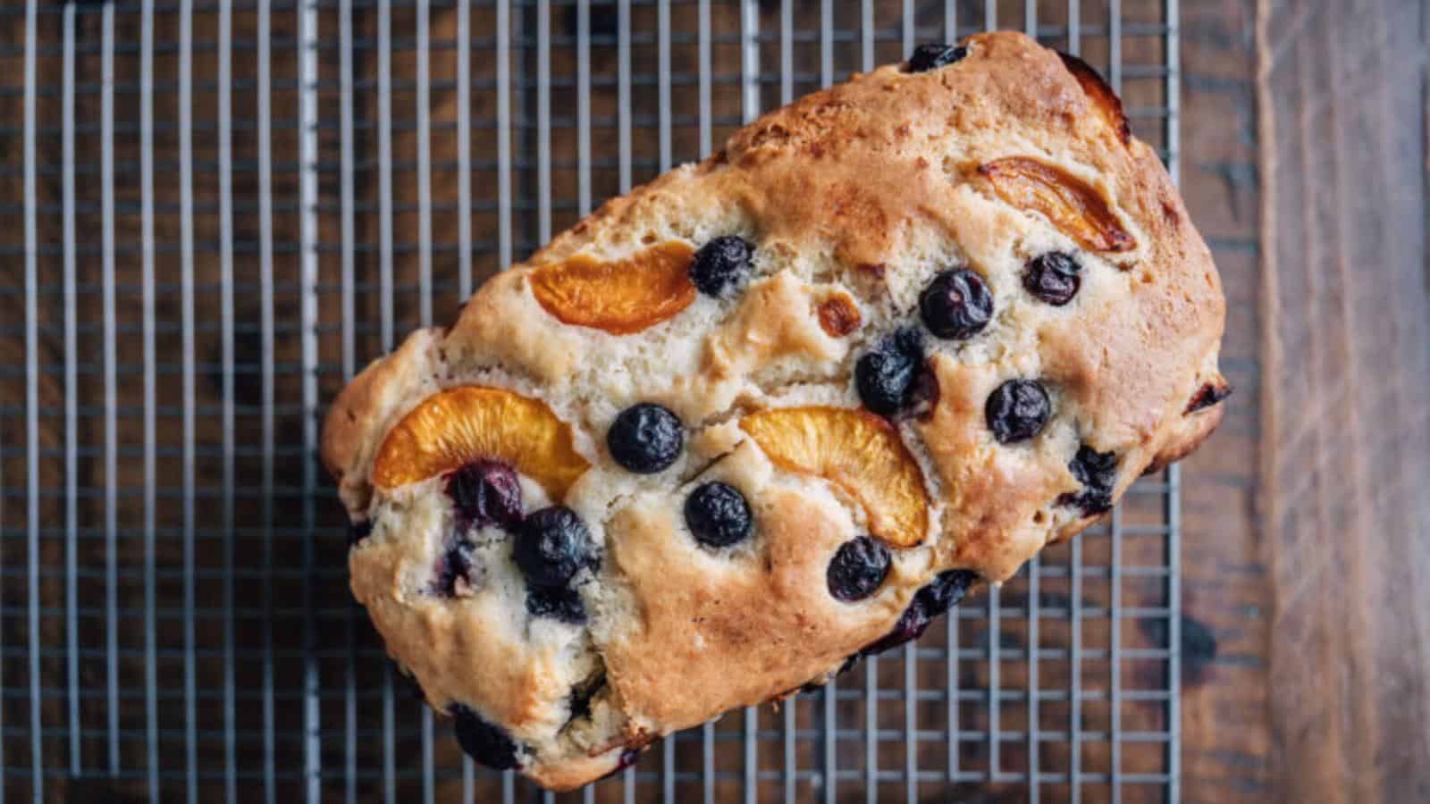 A loaf of fruit bread with blueberries and peach slices cooling on a metal wire rack.