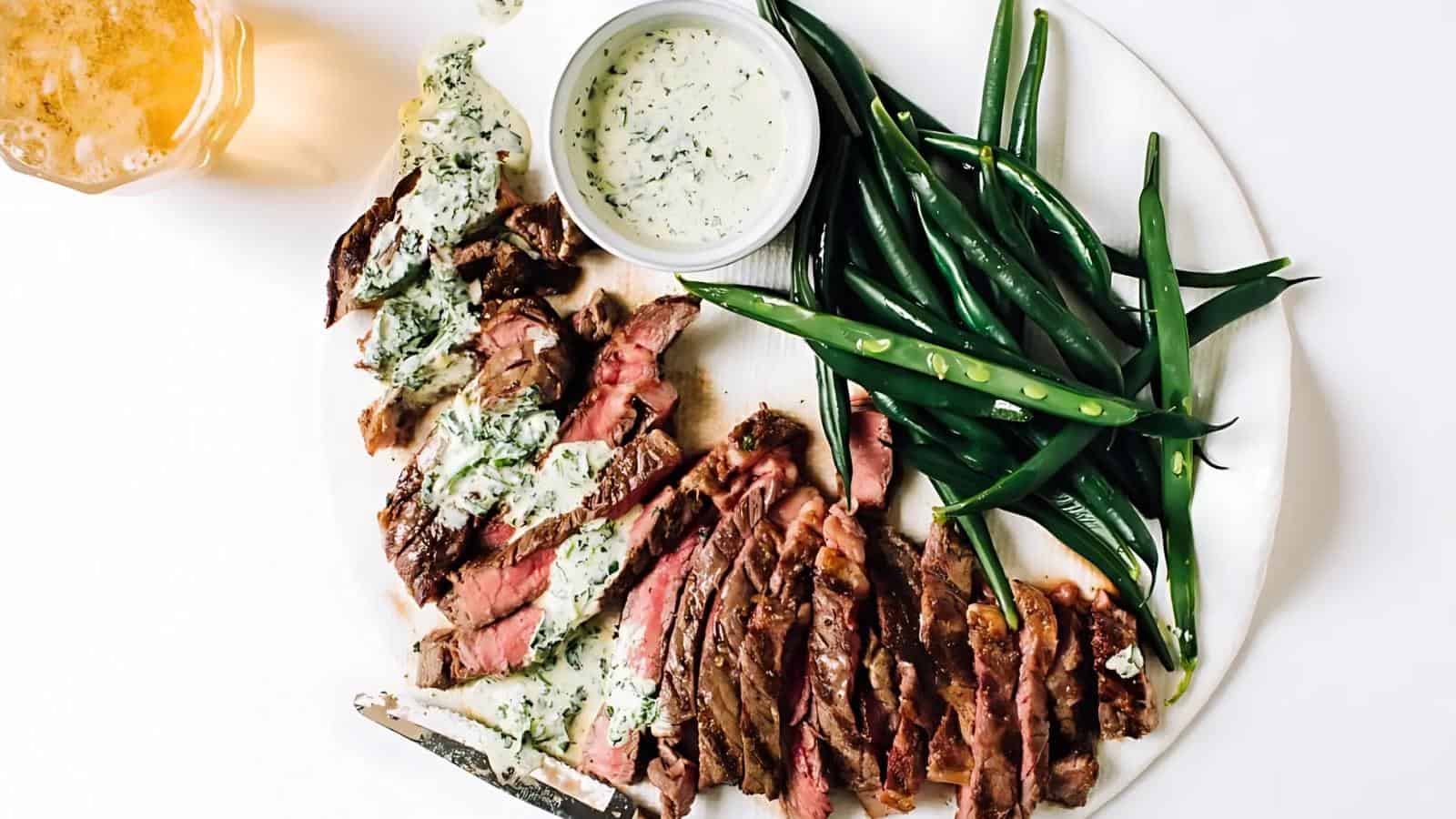 A plate with sliced steak topped with herb sauce, fresh green beans, a small bowl of sauce, and a drink on the side.