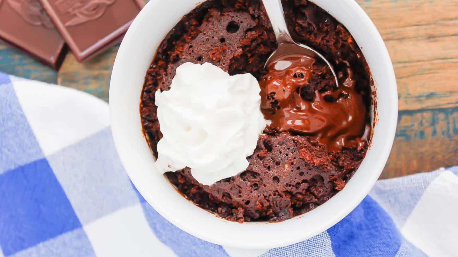 A white mug filled with chocolate mug cake, topped with whipped cream and a spoon, sits on a blue and white checkered cloth.