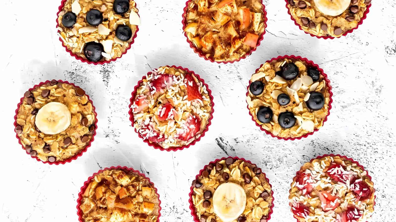 Nine baked oatmeal cups topped with various ingredients including banana slices, blueberries, strawberries, coconut, nuts, and chocolate chips, arranged on a white textured surface.