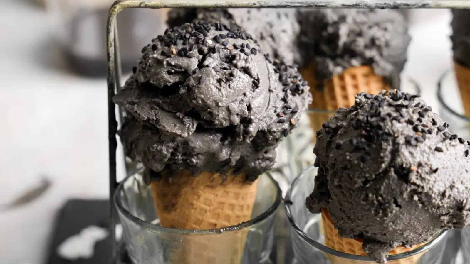 Several waffle cones filled with scoops of black ice cream topped with black sprinkles are arranged upright in clear glasses.