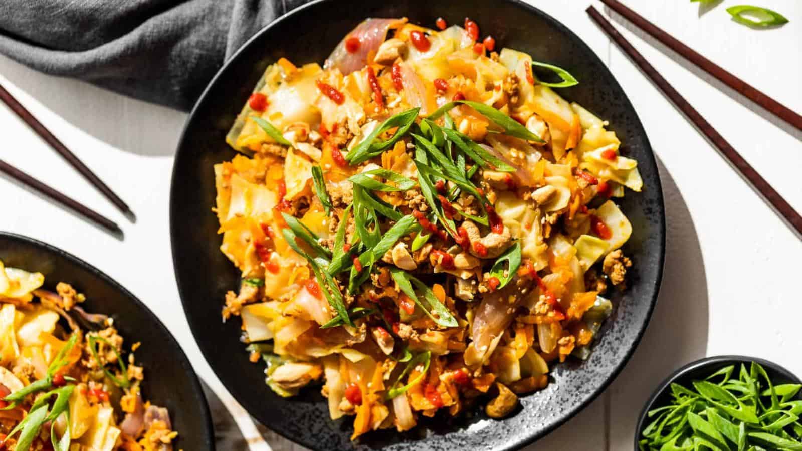 A black plate filled with stir-fried cabbage, noodles, ground meat, sliced green onions, and a drizzle of red sauce, with chopsticks and extra garnishes on the side.