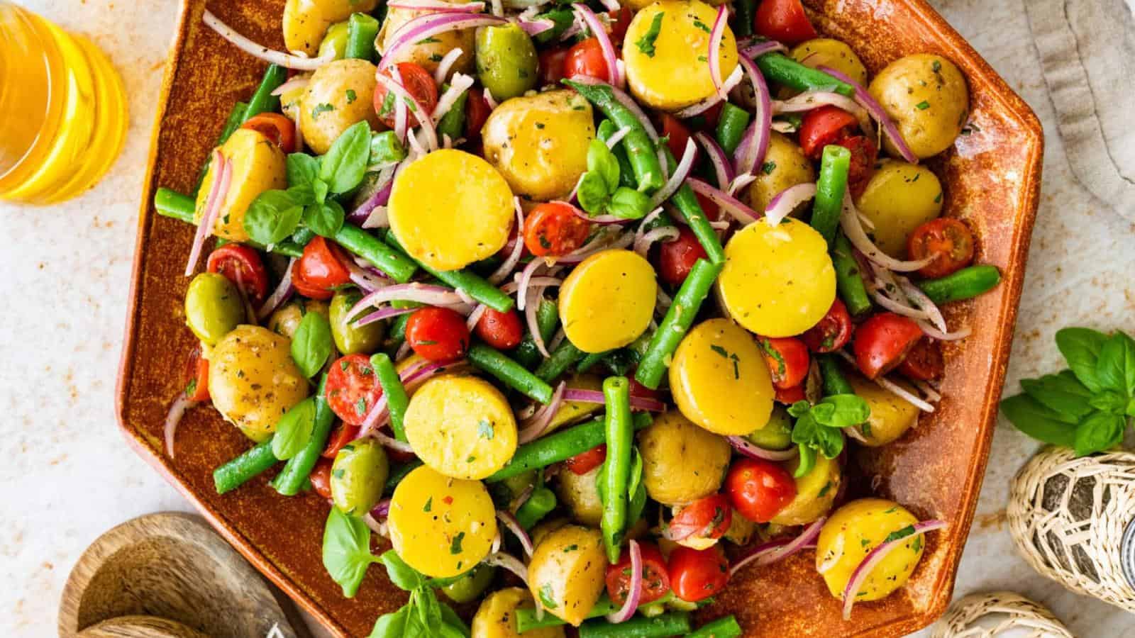 A plate of potato salad with green beans, cherry tomatoes, red onion, green olives, and fresh herbs, dressed in vinaigrette.