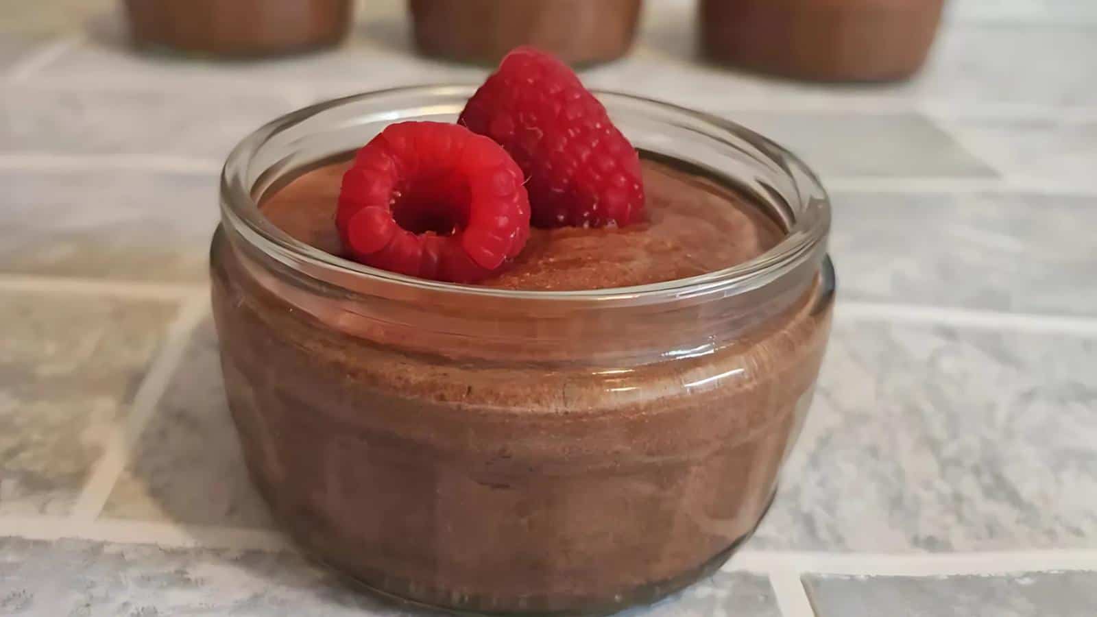 A small glass dish filled with chocolate mousse, topped with two fresh raspberries, sits on a tiled surface.