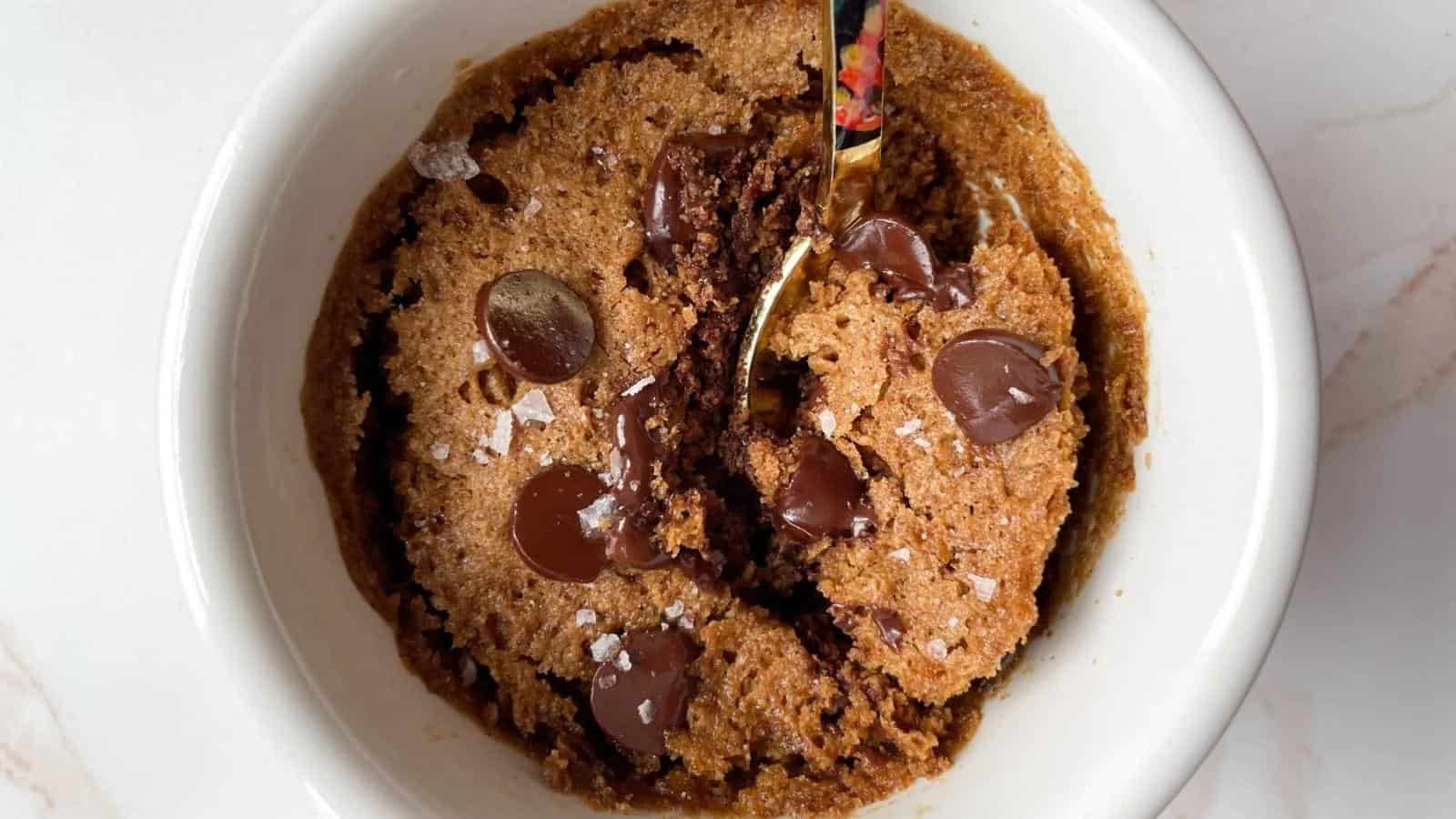 A chocolate chip cookie mug cake with melted chocolate chips and a sprinkling of sea salt, partially scooped with a spoon in a white ramekin.