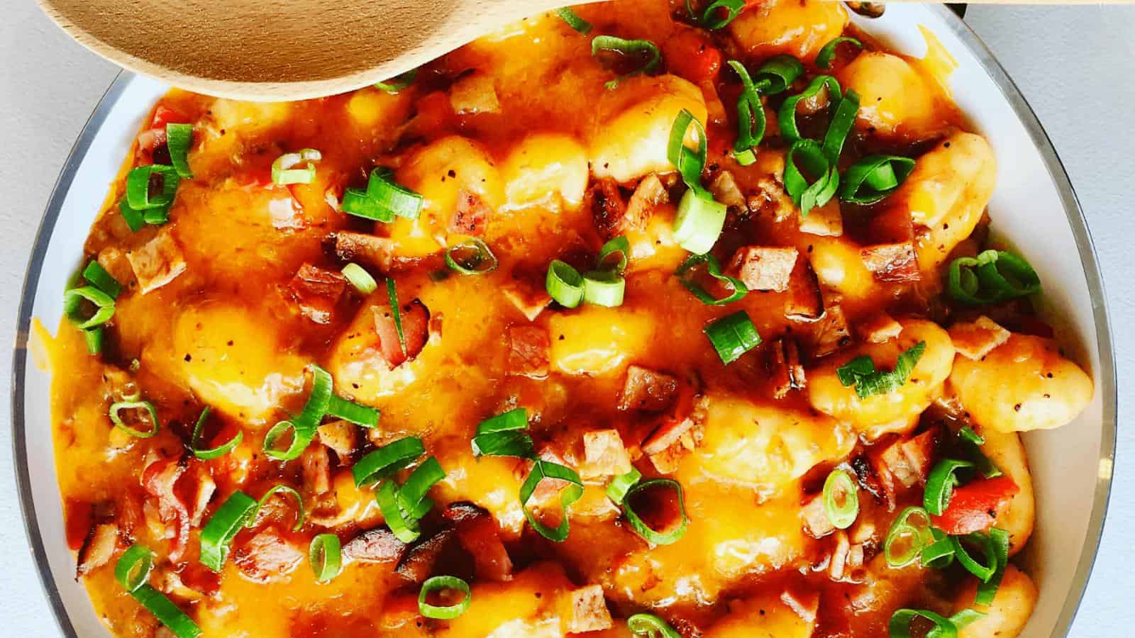 A plate of gnocchi topped with melted cheddar cheese, diced bacon, and chopped green onions, with a wooden spoon resting at the top.