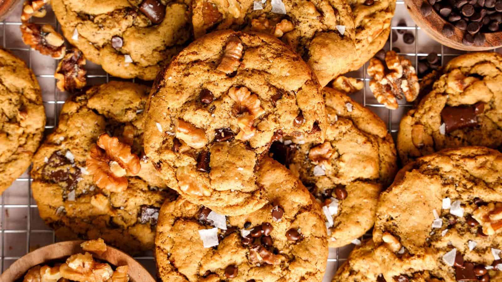 Chocolate chip cookies with walnut pieces are arranged on a cooling rack, with some loose walnuts and chocolate chips visible nearby.