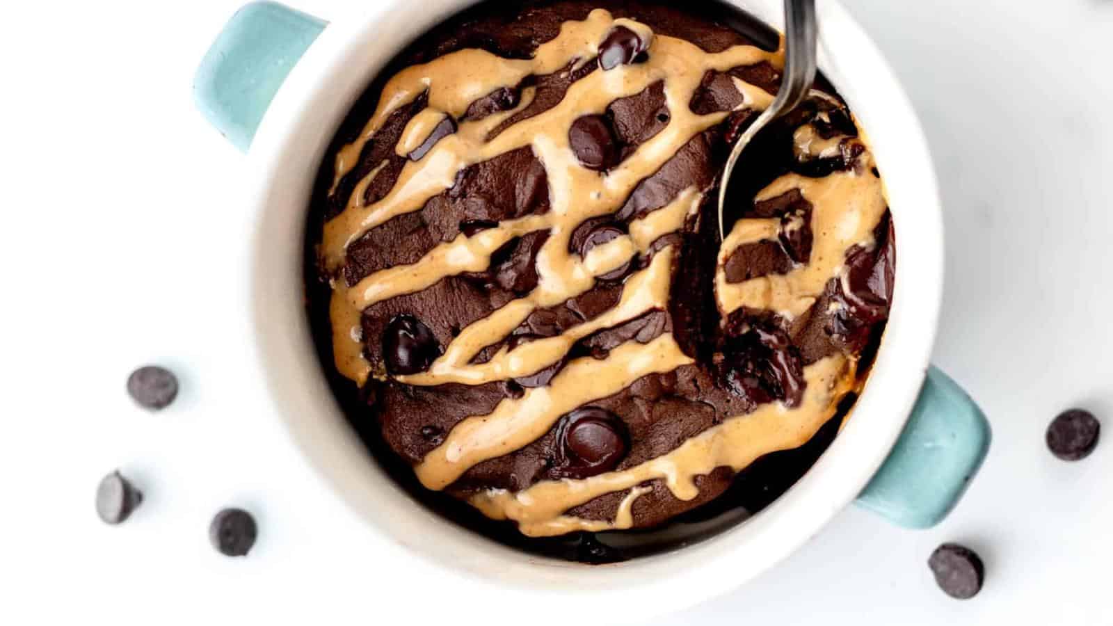 A chocolate mug cake topped with chocolate chips and drizzled with peanut butter, served in a white dish with blue handles and a spoon.