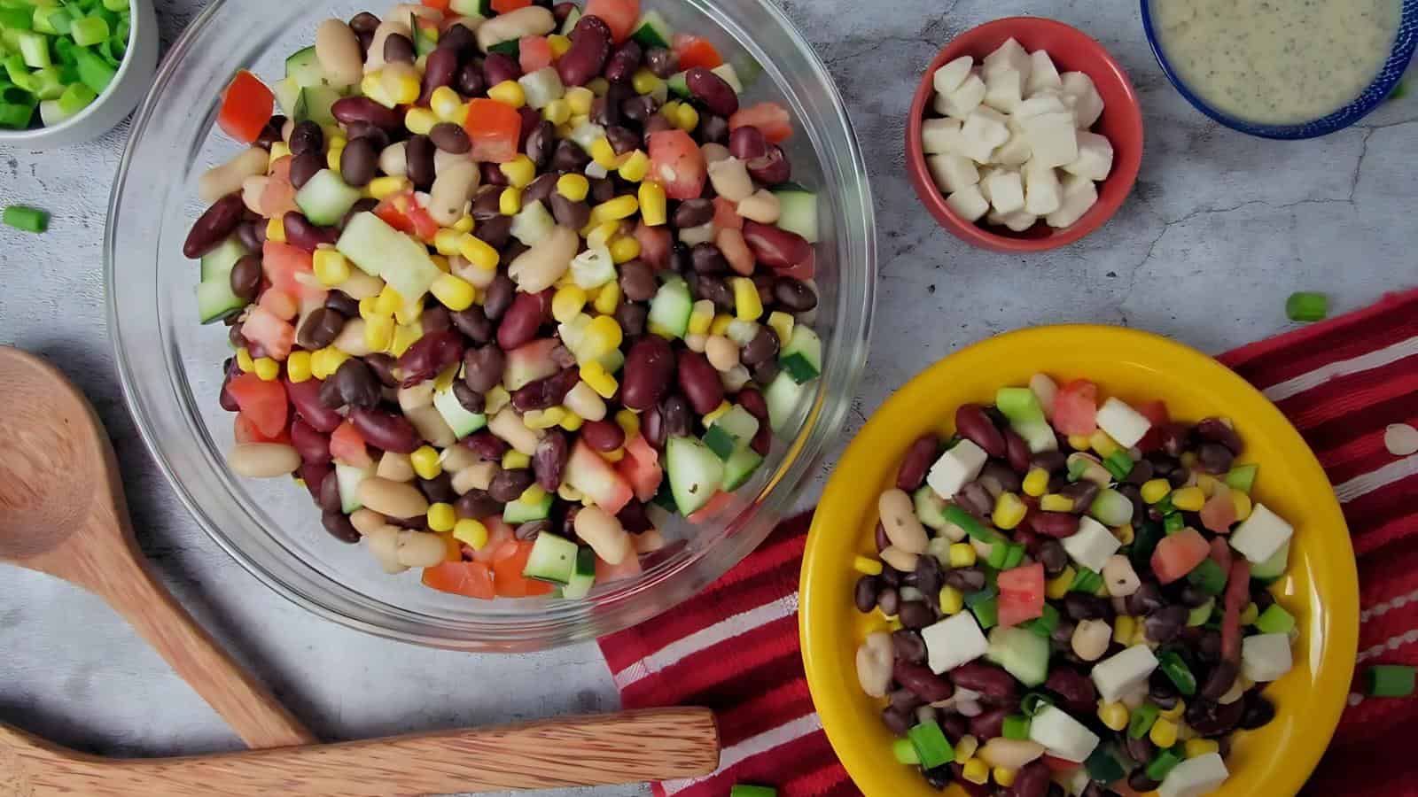 A large glass bowl and a yellow plate filled with a mixed bean salad containing corn, cucumbers, tomatoes, and cheese cubes. Small bowls of diced cheese and green onions are nearby.