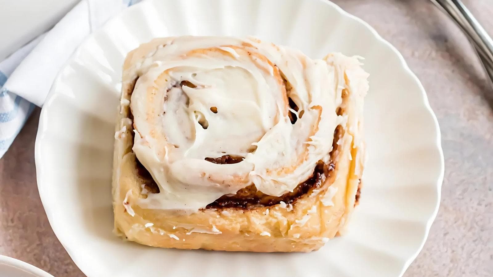 A single cinnamon roll with white icing on top sits on a white plate.