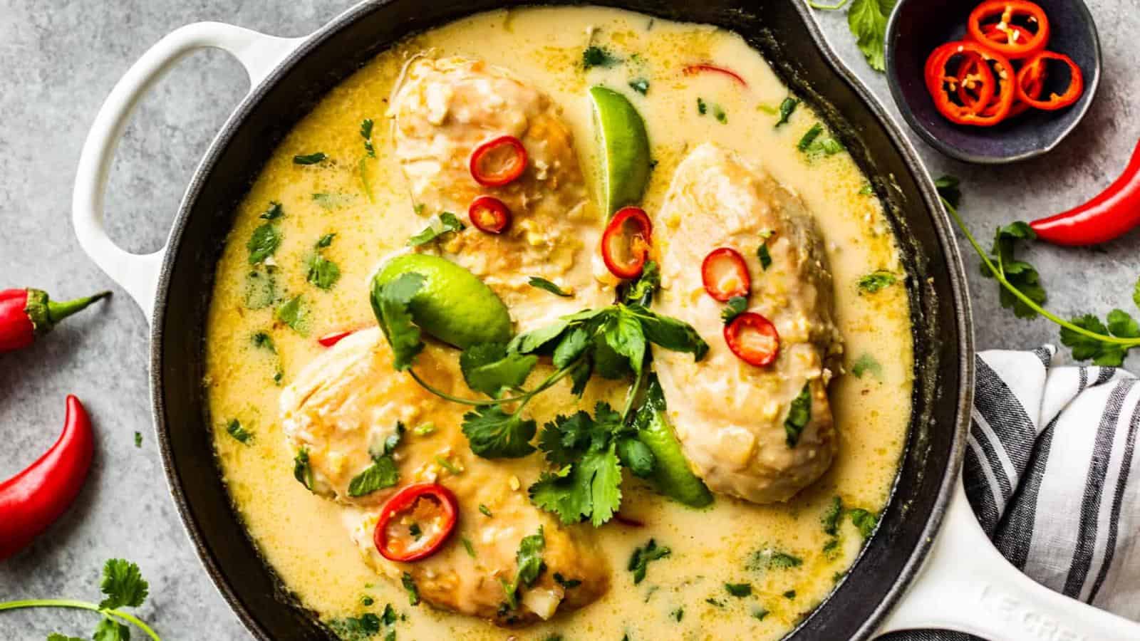 Three cooked chicken breasts in a creamy yellow sauce, garnished with sliced red chili, lime wedges, and cilantro in a white skillet.
