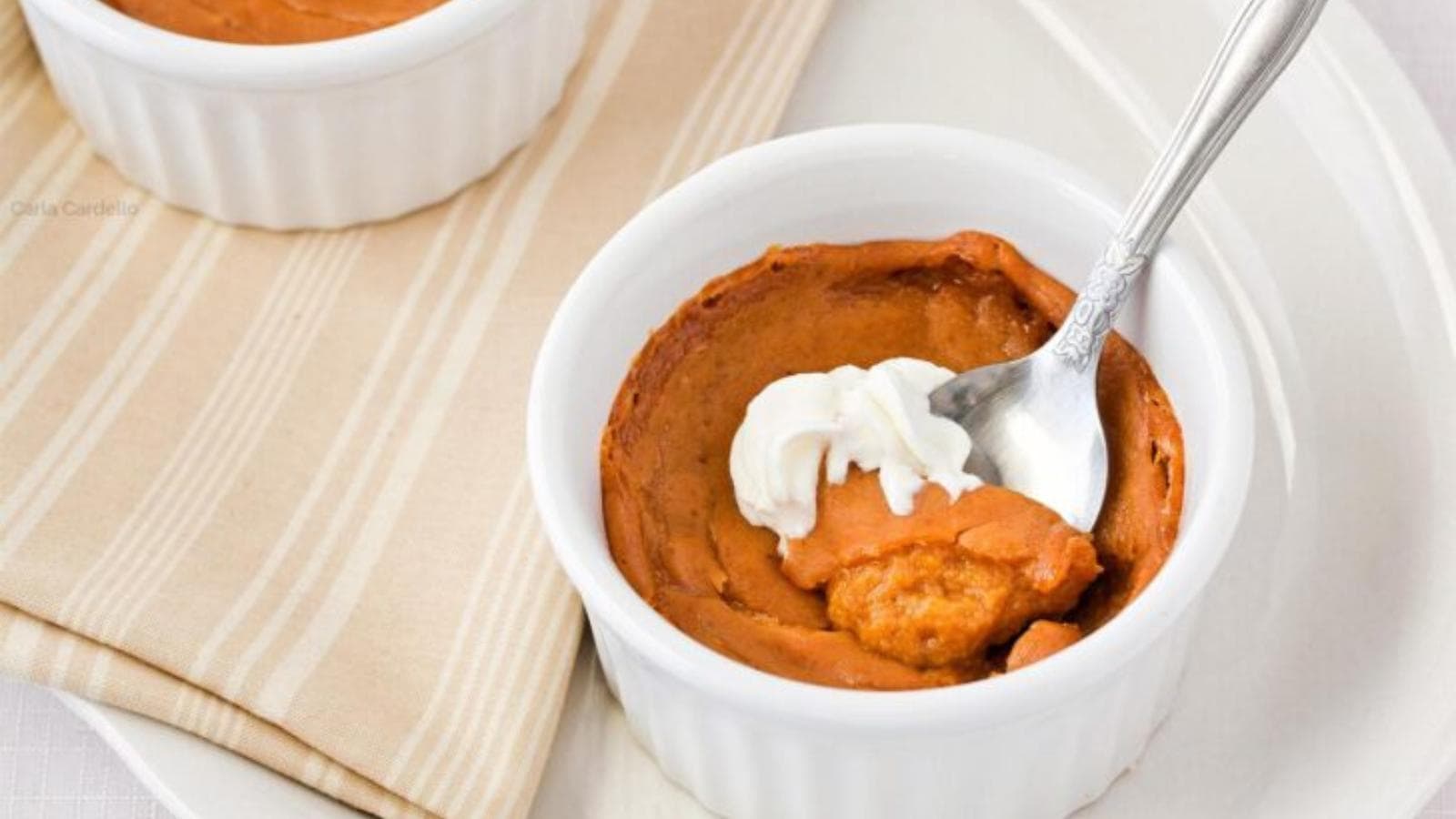 A white ramekin filled with pumpkin pie filling topped with a dollop of whipped cream, with a spoon partially inserted, sits on a white plate next to another ramekin.
