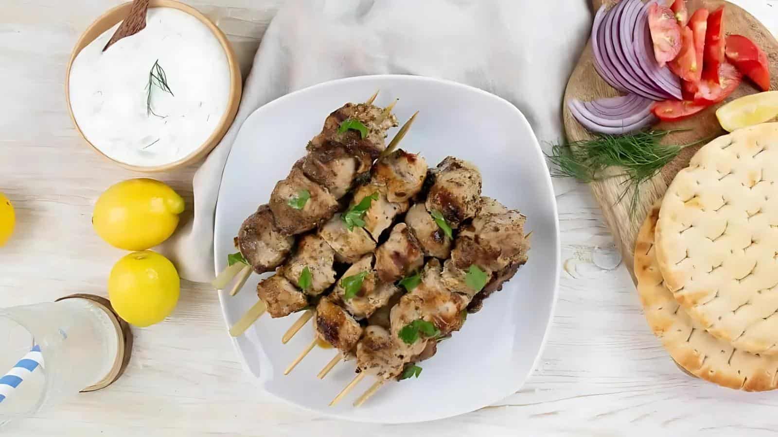 Plate of grilled meat skewers garnished with herbs, surrounded by pita bread, sliced tomatoes, red onions, lemons, and a bowl of white sauce on a wooden table.
