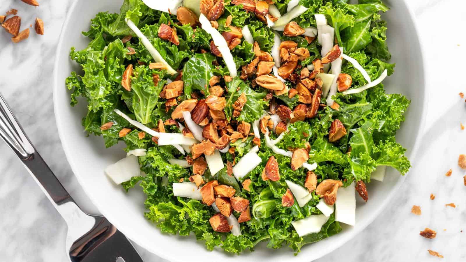 A white bowl filled with chopped kale, sliced almonds, and thin strips of cheese on a marble surface, with salad tongs beside it.
