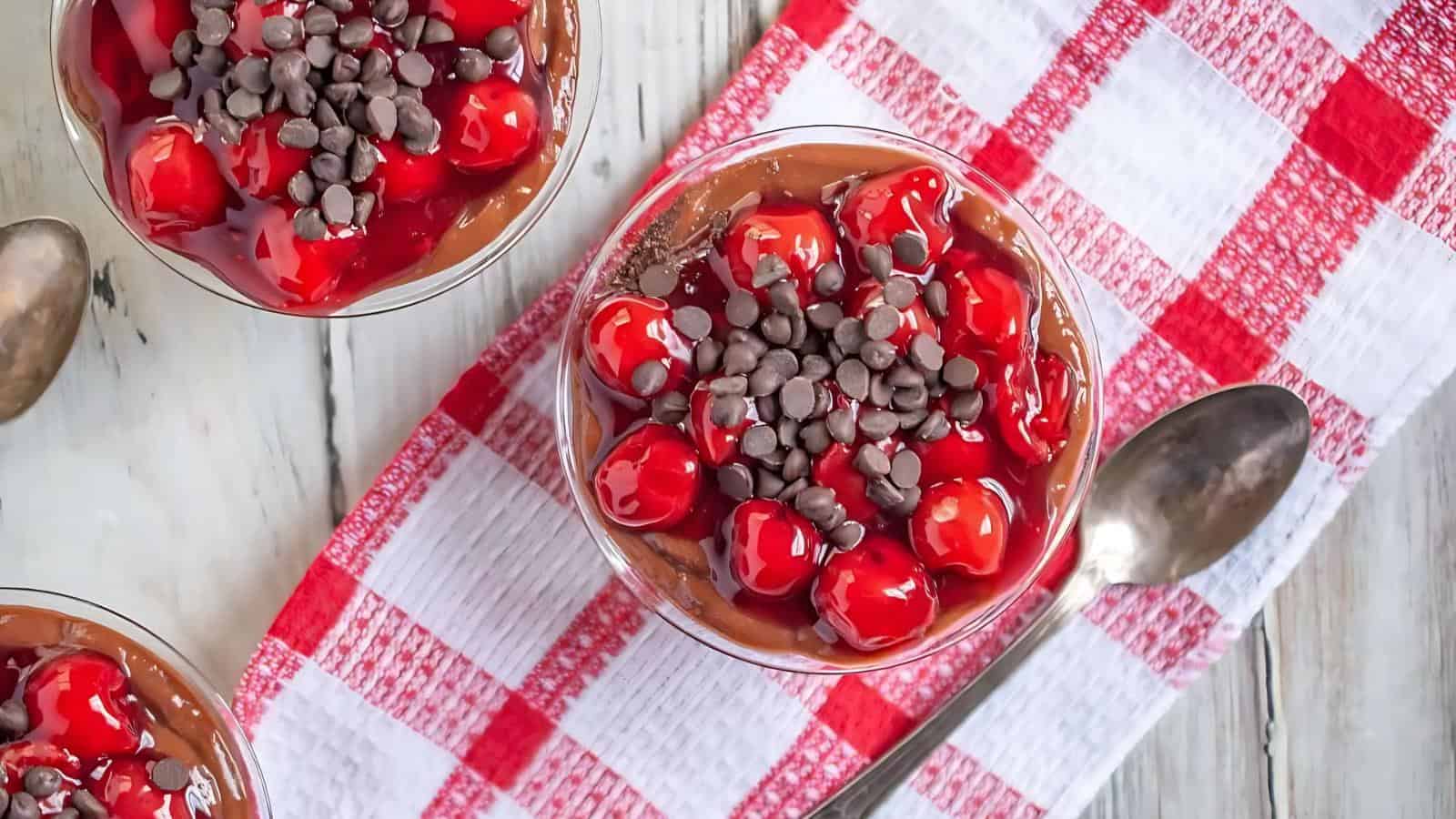 A glass bowl of chocolate pudding topped with cherries and chocolate chips, next to a spoon on a red and white checkered cloth.