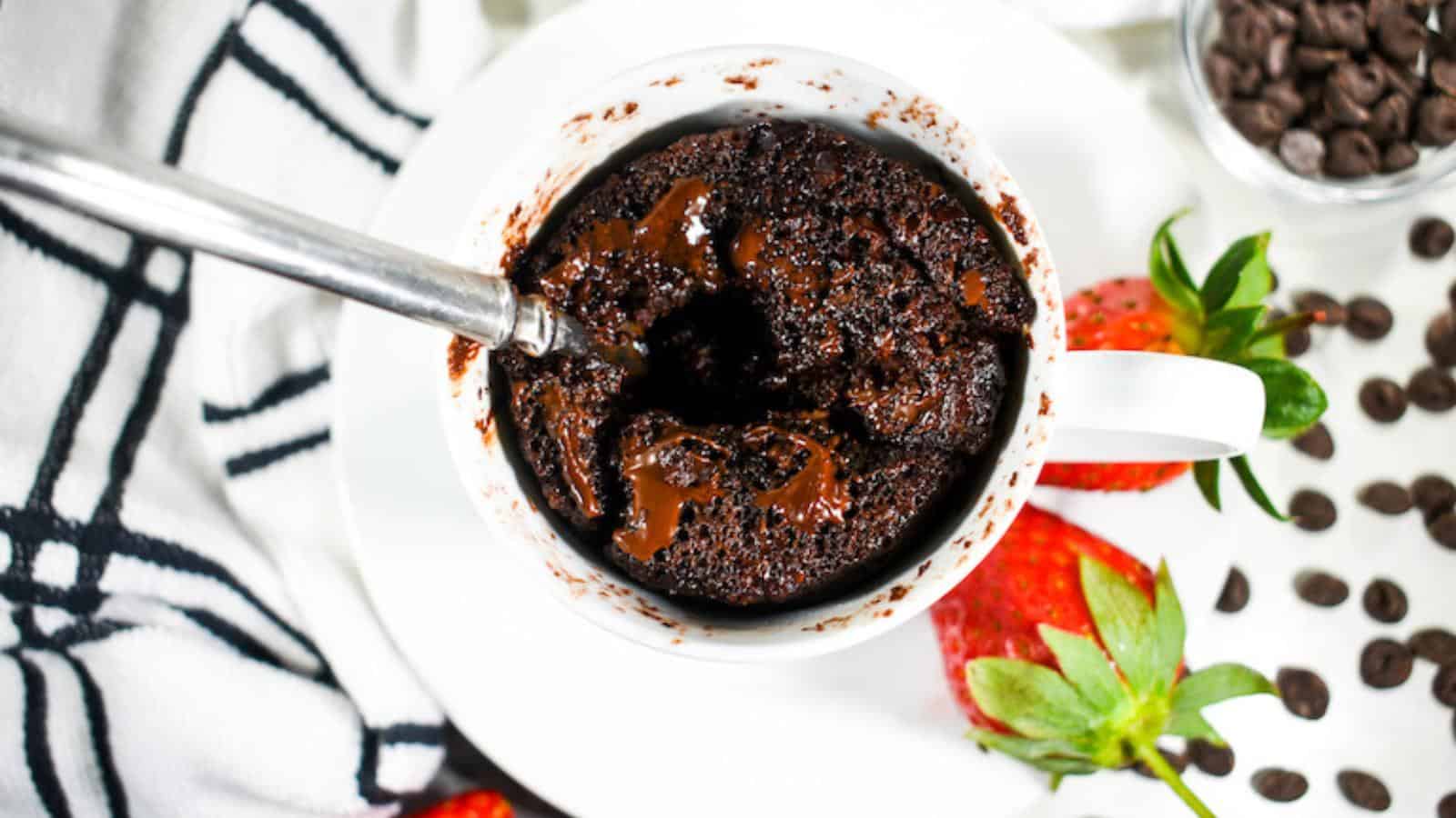 A spoon in a chocolate mug cake with melted chocolate in a white cup, surrounded by strawberries and chocolate chips on a white surface.