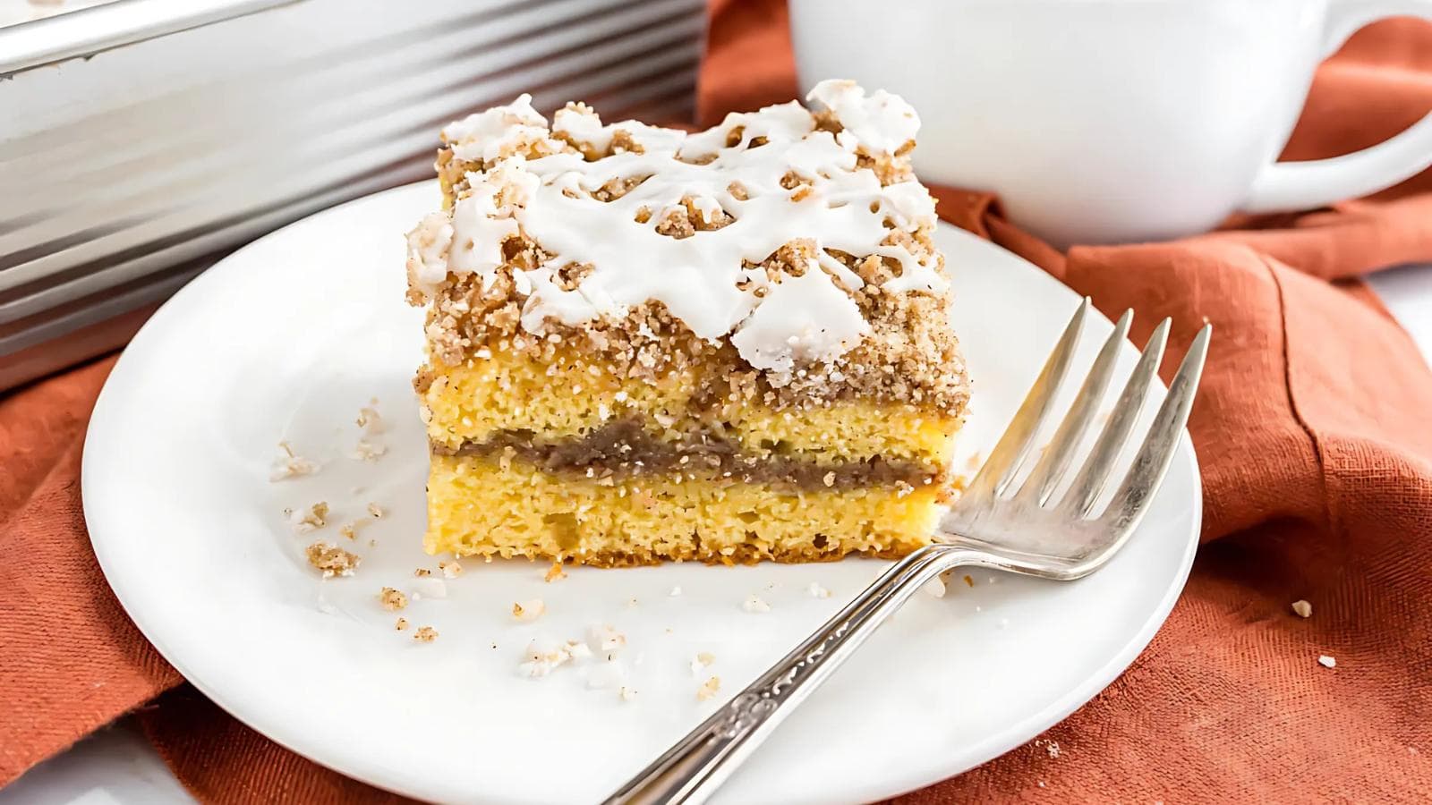A slice of coffee cake with icing and crumb topping sits on a white plate with a fork, on an orange napkin.